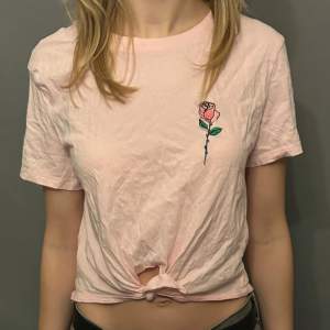 Säljer en söt rosa t-shirt med en broderad ros på bröstet. T-shirten har korta ärmar och en knytning framtill för en trendig look. Perfekt för en avslappnad stil.
