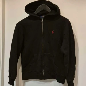 Svart hoodie från Ralph Lauren - Säljer en stilren svart hoodie från Ralph Lauren med dragkedja och det klassiska röda logotypbroderiet på bröstet. Perfekt för en avslappnad look. Den har långa ärmar och en praktisk ficka framtill. Storlek är 14-16 och L