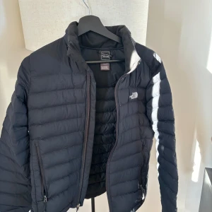 Svart dunjacka från The North Face - Säljer en svart dunjacka från The North Face. Jackan har en dragkedja framtill och två sidofickor med dragkedjor. The North Face-logga på bröstet och ryggen. 