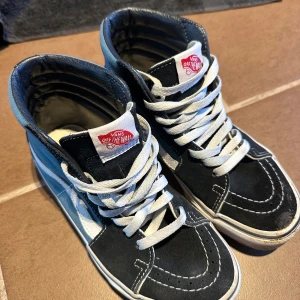 Svarta och blå sneakers från Vans storlek 37 - Säljer ett par klassiska Vans sneakers i svart och blått med vit sula och snörning. Skorna har den ikoniska 'Off the Wall' loggan på plösen och är perfekta för en avslappnad stil.