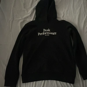 Svart hoodie från Peak Performance - Säljer en stilren svart hoodie från Peak Performance med logga på bröstet. Perfekt för en avslappnad stil. Den har långa ärmar och en klassisk huva.
