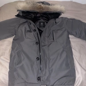 Grå parkajacka från Canada Goose - Säljer en grå parkajacka från Canada Goose med pälskantad huva. Jackan har dragkedja och knappar framtill samt flera praktiska fickor. Perfekt för kalla vinterdagar. 🧥❄️