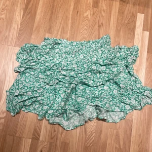 Grön blommig kjol från Samsa Collection - Charmig grön kjol med blommigt mönster från Samsa Collection. Kjolen har volanger som ger en lekfull känsla och är perfekt för varmare dagar. Den elastiska midjan gör den bekväm att bära. Säljer pga att den inte används