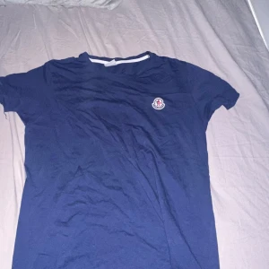 Blå t-shirt från Moncler - Säljer en snygg blå t-shirt från Moncler med korta ärmar och en liten logga på bröstet. Perfekt för en avslappnad stil. T-shirten har en klassisk rund halsringning med vit kant.