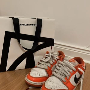Nike Dunk i orange och vit - Snygga Nike Dunk sneakers i en fräsch kombination av orange och vit med svarta detaljer. Skorna har klassisk snörning och en bekväm passform. Perfekta för en sportig look. Storlek 37