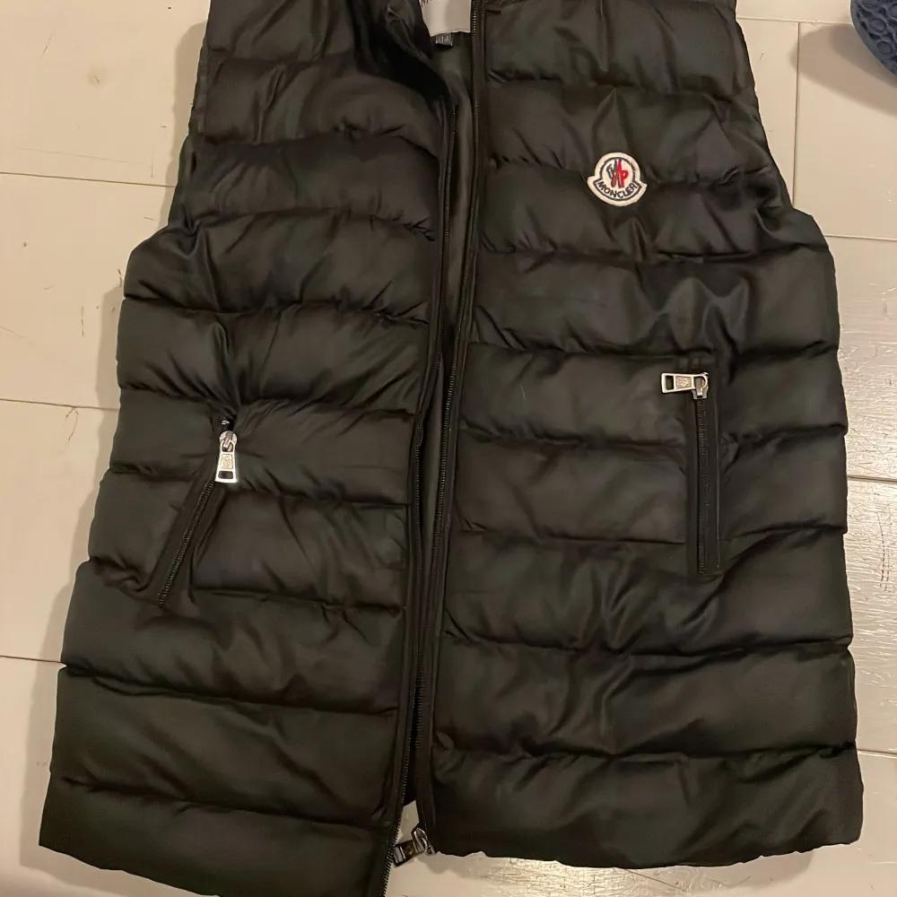 Snygg svart dunväst från Moncler med dragkedja och logotyp på bröstet. Västen har två praktiska fickor med dragkedjor framtill. !Inte äkta!. Takit.