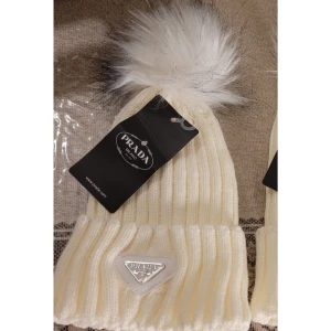 Vit ribbad mössa med pälsboll från Prada - Snygg vit ribbad mössa från Prada med en fluffig pälsboll på toppen. Mössan har en elegant design med en metallogo framtill. Perfekt för att hålla sig varm och stilren under kalla dagar.