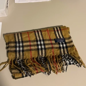 Burberry Check Scarf - Säljer en klassisk rutig halsduk från Burberry i beige, svart, vit och röd. Tillverkad i en lyxig blandning av merino, angora och kashmir. Perfekt för att hålla dig varm och stilren under kyliga dagar. Fransen ger en extra touch av elegans.