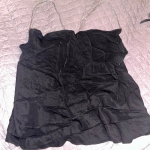 Svart linne med kedjeband - Snyggt svart linne med tunna kedjeband som ger en elegant touch. Perfekt för en kväll ute eller en speciell tillställning. Materialet är mjukt och följsamt.