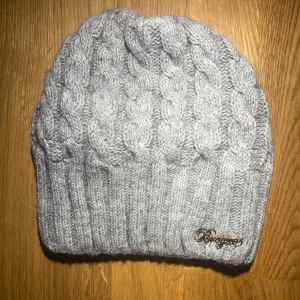 Grå stickad mössa  - Snygg grå stickad mössa  med flätmönster och ribbad kant. Perfekt för kyliga dagar och håller dig varm och stilren. Den har en diskret logga i metall på sidan.🤍