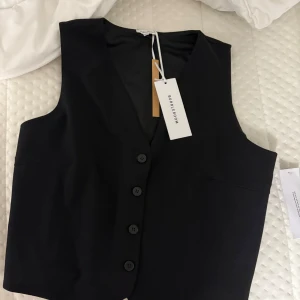 Svart väst från Bubbleroom - Snygg svart väst från Bubbleroom med knappar framtill. Perfekt för att ge en stilren touch till din outfit. Västen har en klassisk design och är lätt att matcha med olika plagg.