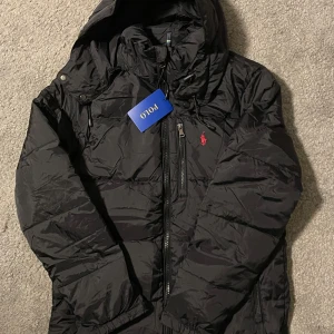Polo Ralph Lauren jacka - Säljer en svart dunjacka från Polo med huva och dragkedja. Jackan har en broderad röd logotyp på bröstet och är perfekt för kyligare väder. Den är långärmad och har en stilren design. Storlek M (Helt ny)