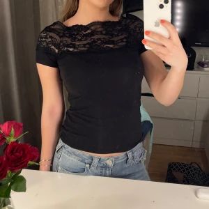 Svart topp med spetsdetaljer - Säljer en svart topp med vacker spetsdetalj vid halsringningen. Toppen har korta ärmar och en tight passform, perfekt för en stilren look. Det finns en liten defekt på tyget som syns på bilden. Det är ett litet hål, men väldigt litet. Vet inte märket då lappen är bortklippt. 