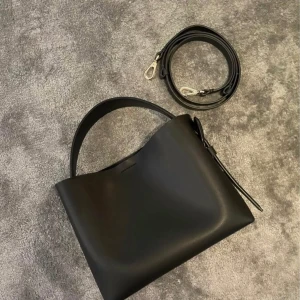 ARKET Svart axelväska med avtagbar rem  - Snygg svart axelväska i skinn med avtagbar axelrem. Väskan har en stilren design och är perfekt för att bära dina dagliga nödvändigheter. Den har en enkel och elegant look som passar till många olika stilar. Mått  23 cm x 19 cm x 10,5 cm.