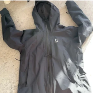 Svart vindjacka från Haglöfs - Säljer en svart vindjacka från Haglöfs med huva och dragkedja. Jackan har långa ärmar och flera praktiska fickor. Perfekt för utomhusaktiviteter och skyddar mot vind och regn.