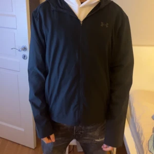 Svart träningsjacka från Under Armour - Snygg svart träningsjacka från Under Armour med huva och dragkedja. Ej använd, säljer eftersom den är för stor för mig! 