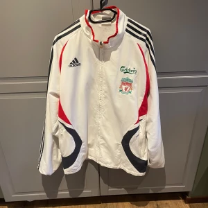 Vit vindjacka från Adidas med Liverpool FC-logga - Säljer en vit vindjacka från Adidas med Liverpool FC-logga och Carlsberg-tryck. Jackan har svarta och röda detaljer samt dragkedja framtill. Perfekt för Liverpool supporter.