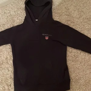 Mörkblå  hoodie från GANT - Säljer en stilren svart hoodie från GANT med logotyp på bröstet. Tröjan har en klassisk design med huva och en praktisk magficka. 