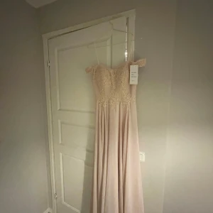 Champagnefärgad långklänning med spetsdetaljer - Elegant champagnefärgad långklänning med vackra spetsdetaljer och offshoulder-design. Klänningen har en romantisk känsla med sin flytande kjol och detaljerade överdel. Perfekt för speciella tillfällen.