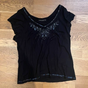 Svart topp med paljettdetaljer från Twilfit - Snygg svart topp från Twilfit med v-ringning och glittrande paljettdetaljer i blommönster. Toppen har korta ärmar och en elegant design som passar perfekt för en kväll ute. Perfekt för dig som vill ha något unikt och stilrent i garderoben.