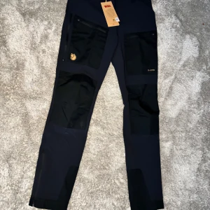 Svarta friluftsbyxor från Fjällräven - Snygga och funktionella svarta friluftsbyxor från Fjällräven med förstärkta knän och flera praktiska fickor. Byxorna har en hög midja och är perfekta för utomhusaktiviteter. Tillverkade i slitstarkt G-1000 material för extra hållbarhet.