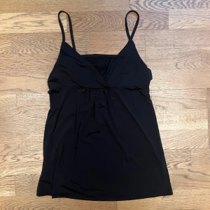 Svart omlottlinne med spetsdetalj - Snyggt svart linne med smala axelband och omlottdesign framtill. Linnet har en subtil spetsdetalj vid bysten som ger en elegant touch. Perfekt för en stilren look.