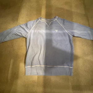 Blå sweatshirt från GANT - Säljer en stilren blå sweatshirt från GANT med vita sömmar och liten logga på bröstet. Tröjan har långa ärmar och en klassisk rund halsringning. Perfekt för en avslappnad stil.