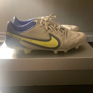  fotbollsskor - Snygga Nike Tiempo fotbollsskor i grått med gula och blå detaljer. Skorna har snörning och är perfekta för fotbollsplanen. De har en klassisk design med Nike-loggan på sidan.