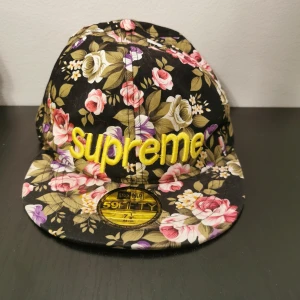 Blommig keps  - Snygg keps köpt utomlands  med ett färgglatt blommönster på svart bakgrund. Kepsen har broderad text i gult framtill och är perfekt för att ge din outfit en unik touch.