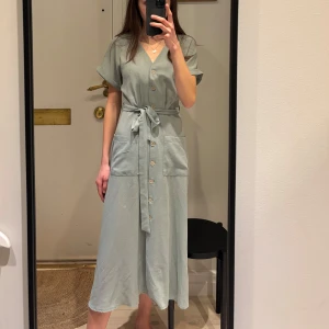 Mintgrön klänning med knappar och knytband - Snygg grön/blå klänning med v-ringning och knappar framtill. Den har korta ärmar och ett knytband i midjan för en smickrande passform. Perfekt för en avslappnad stil.