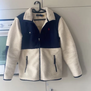 Beige och marinblå teddyjacka från Ralph Lauren - Säljer en snygg teddyjacka från Ralph Lauren i vit och marinblått. Jackan har en dragkedja framtill och två fickor med dragkedja. Den har en klassisk krage och en liten röd logga på bröstet. Perfekt för kyliga dagar!