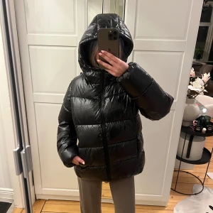 Svart pufferjacka med huva - Oanvänd glansig jacka i storlek 158/164, passar mig som är en xxs-xs. Öppna fickor framtill. Perfekt för vintern. Köptes för 800kr på KidsBrandStore. Säljer ej för lägre pris.