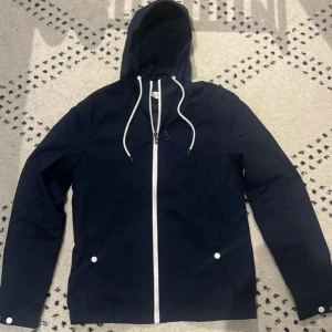 Mörkblå vindjacka med huva - Säljer en stilren mörkblå vindjacka med vit dragkedja och snören i huvan. Jackan har långa ärmar och två fickor med knappar framtill. Perfekt för blåsiga dagar.