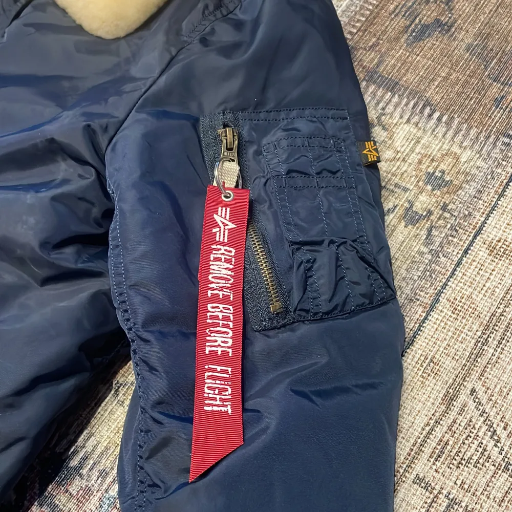 Säljer en snygg blå bomberjacka från Alpha Industries med en mjuk, beige pälsliknande krage. Jackan har en dragkedja framtill och en cool detalj med 'Remove Before Flight'-tag på ärmen. Perfekt för höst och vinter!. Takit.