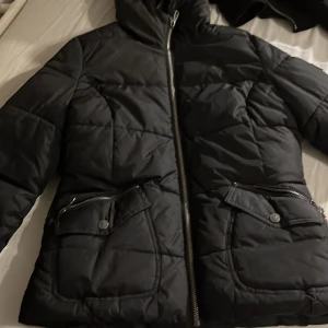 Svart dunjacka från Navahoo - Säljer en svart dunjacka från Navahoo med mysigt vitt foder. Jackan har en dragkedja framtill och praktiska fickor med knappar. Perfekt för kalla dagar med sin quiltade design och varma känsla.