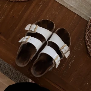 Birkenstock - Säljer ett par klassiska vita sandaler från Birkenstock med två justerbara remmar och spännen i metall. Sandalerna har en bekväm korksula som formar sig efter foten för extra komfort.