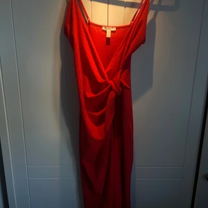 Röd klänning med slit där fram - Snygg röd klänning med en v-ringad design och knytdetalj och slit framtill. Klänningen har smala axelband och är djup v ringad . 