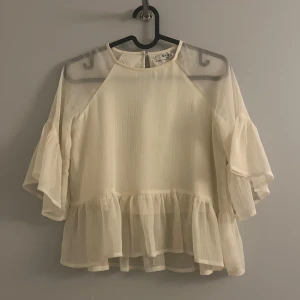 Beige/ljus gul blus med volangdetaljer - Säljer en söt beige/ljus gul blus från NA-KD med volangdetaljer och trekvartsärmar. Blusen har en luftig och lätt design, perfekt för varmare dagar. Den är tillverkad i ett transparent material som ger en elegant look.
