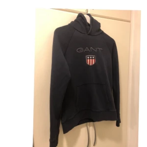 Mörkblå hoodie från GANT strl 176 - Säljer en snygg mörkblå hoodie från GANT med broderad logga och emblem på bröstet. Tröjan har en klassisk känguruficka och en bekväm huva. Perfekt för en avslappnad stil. Kommer från djur och rökfritt hem 