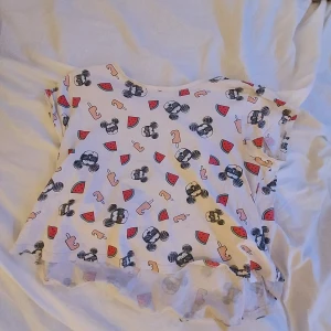 Vit t-shirt med Mussepigg-mönster - Säljer en vit t-shirt från Disney med ett lekfullt mönster av vattenmeloner, solglasögon och musseöron. T-shirten har korta ärmar och en rund halsringning.