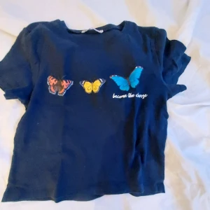 Mörkblå t-shirt med fjärilar från FB Sister - Säljer en mörkblå t-shirt från FB Sister med tre färgglada fjärilar och texten 'become the change' på framsidan. T-shirten har korta ärmar och är gjord i bomull. Perfekt för en avslappnad stil.