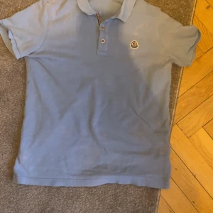 Moncler Piké - Säljer en ljusblå pikétröja från Moncler med korta ärmar och klassisk krage. Tröjan har en broderad Moncler-logga på bröstet och knappar framtill. Perfekt för en avslappnad stil.
