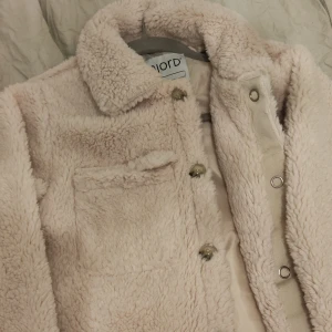 Beige teddyjacka från Njord - Mysig beige teddyjacka från Njord med knappar framtill och en bröstficka. Perfekt för kyliga dagar med sin fluffiga och varma design. Jackan har en klassisk krage och är långärmad.