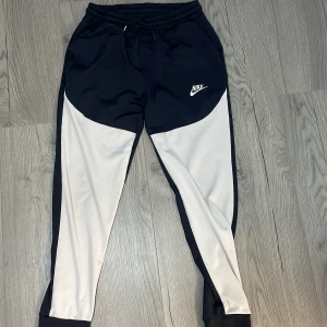 Svarta och vita mjukisar från Nike - Snygga träningsbyxor från Nike i svart och vitt. De har en elastisk midja med snörning och ett broderat Nike-logotyp på benet. Perfekta för träning eller en avslappnad dag.