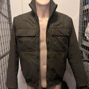 Olivgrön jacka från H&M L.O.G.G. - Säljer en olivgrön jacka från H&M L.O.G.G. med dragkedja och ribbad krage. Jackan har två bröstfickor med lock och är quiltad på insidan för extra värme. Perfekt för kyligare dagar! 