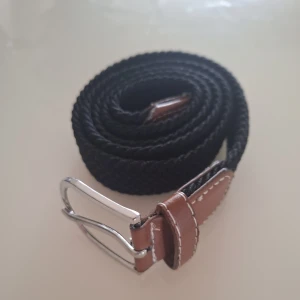 Flätat bälte  - Snyggt svart flätat bälte med bruna skinnliknande detaljer och silverfärgat spänne. Perfekt för att ge en stilren touch till din outfit. Bältet har en klassisk design med kontrasterande sömmar.