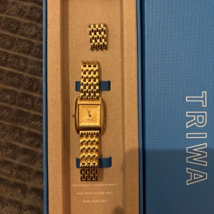 Guldklocka från Triwa - Säljer en stilren Triwa klocka med en vacker guldfärgad design. Klockan har en rektangulär urtavla med Triwas logotyp och en feminin symbol. Armbandet är också guldfärgat och har en klassisk länkdesign. Ett extra armband medföljer.  Använd ett par gånger, väldigt fint skick!  Nypris: 1995:-  Mer info finns på Triwas hemsida