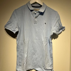 Ljusblå pikétröja - Snygg ljusblå pikétröja med klassisk krage och knappar framtill. Tröjan har korta ärmar och en liten broderad logotyp på bröstet. Perfekt för en avslappnad stil.