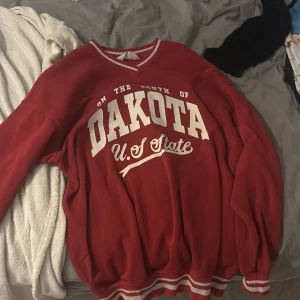 Röd tröja från H&M med tryck - Säljer en röd tröja från H&M med texten 'Dakota U.S State' tryckt på framsidan. Tröjan har långa ärmar och ribbade muddar i rött och vitt vid ärmslut och nederkant.  Har använt 3-4 gånger men gillade inte den