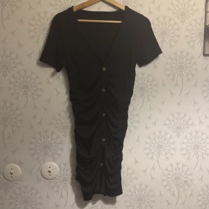 Svart klänning med knappar - Snygg svart klänning med v-ringning och knappar framtill. Den har en åtsittande passform med rynkade detaljer som ger en smickrande siluett. Perfekt för en stilren look.